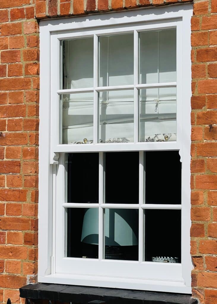 A white-framed timber sash window set in a red brick wall, with the lower sash open. Behind the glass, a partially visible table lamp and some small decorative items can be seen inside the room.