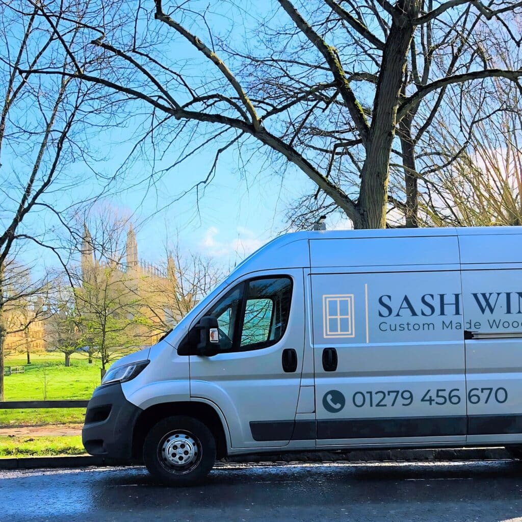 A white van displaying "Sash Windows Custom Made Wood" is parked on a street lined with bare trees. Beyond, a grassy area and a historic building with towers rise against a clear blue sky—ideal surroundings to understand the essence of how to protect your windows and doors effectively.