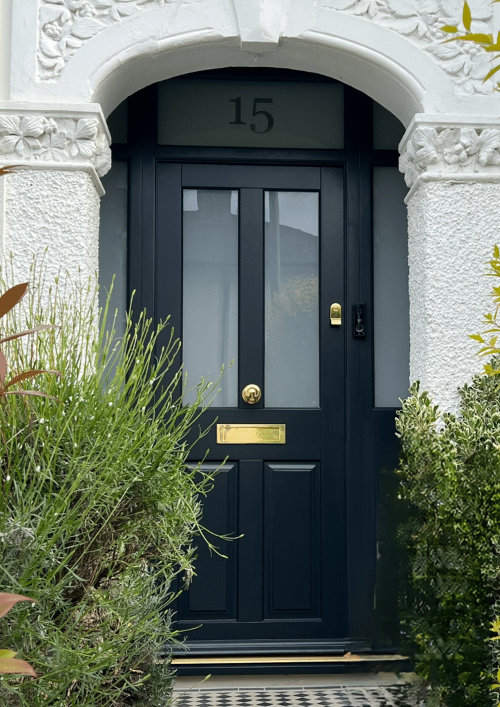 A black wooden door with four glass panels and a brass mail slot stands in a white stone wall adorned with ornate floral carvings, showcasing the charm of timber windows and doors in Cambridge. The house number "15" sits above it, surrounded by lush green bushes.