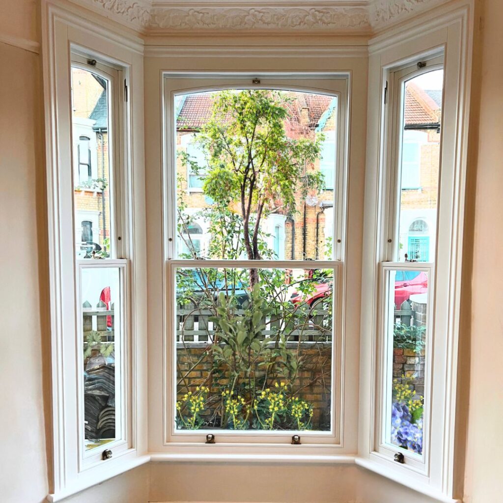 A view through a bay window with three panels, overlooking a small garden. The garden features greenery and yellow flowers, with brick houses visible in the background. The window has a classic white frame and ornate molding.