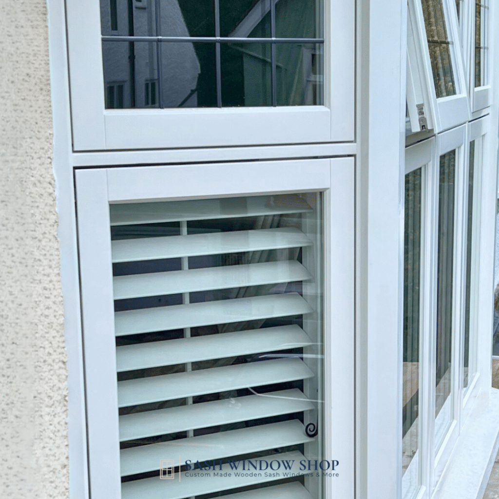 A white framed window with partially open wooden blinds accompanies a sleek set of casement windows, their clear glass panels angled to the side. The frame casts subtle shadows against the building's lightly textured exterior.