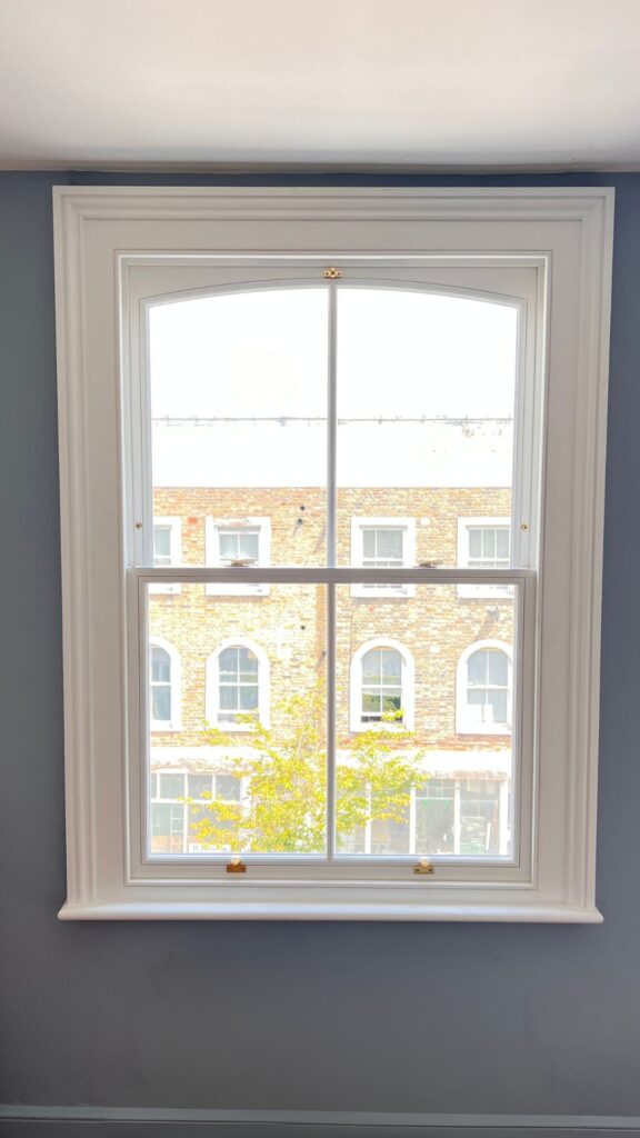 A classic white-framed window with a view of a brick building and a tree outside. Timber sliding sash windows complement the scene, while the wall surrounding the window is painted in a light blue color.