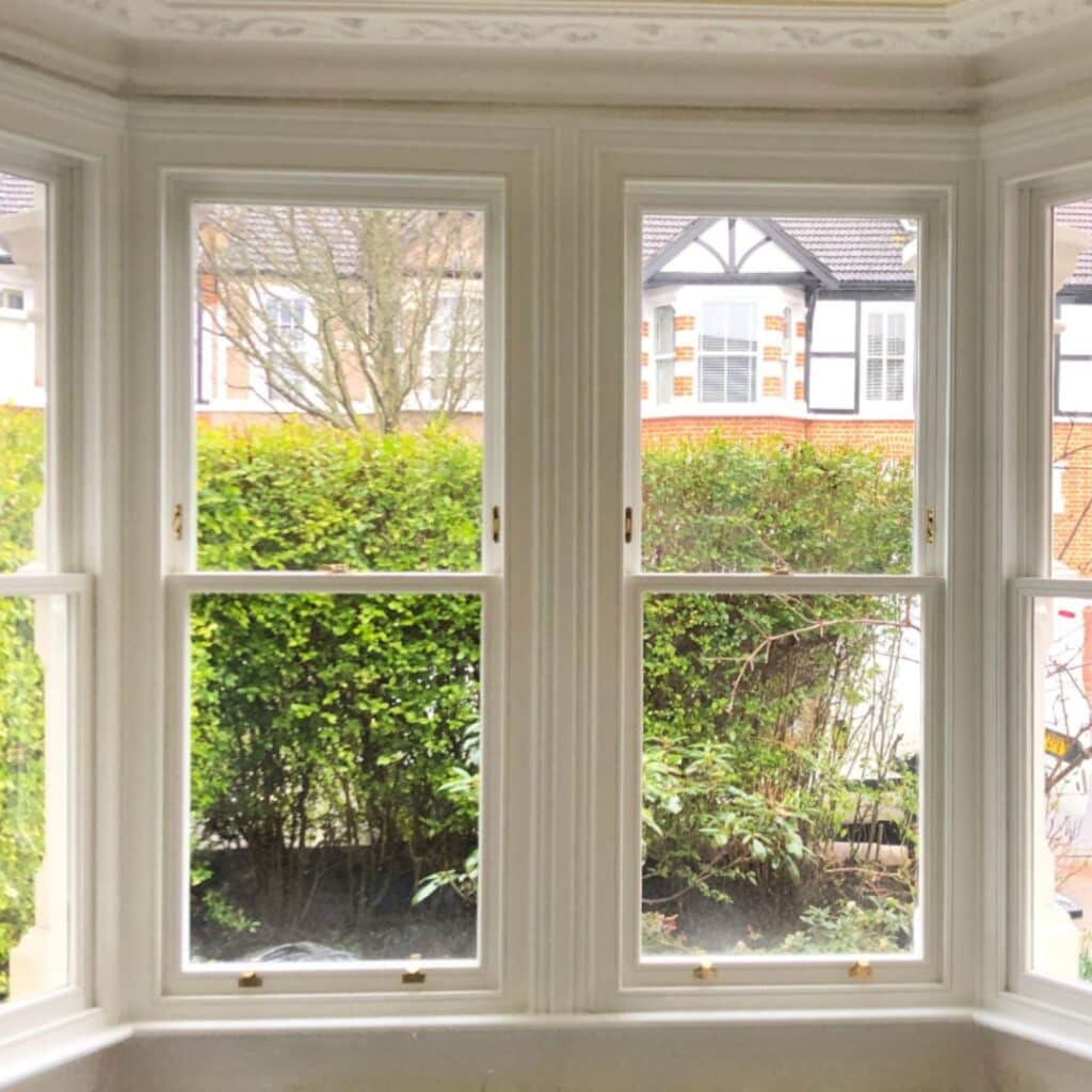 A view from inside a room through three timber sash windows reveals a green garden with bushes and a suburban street with houses. The white-framed windows showcase the bright and clear scene, depicting a peaceful neighborhood setting.