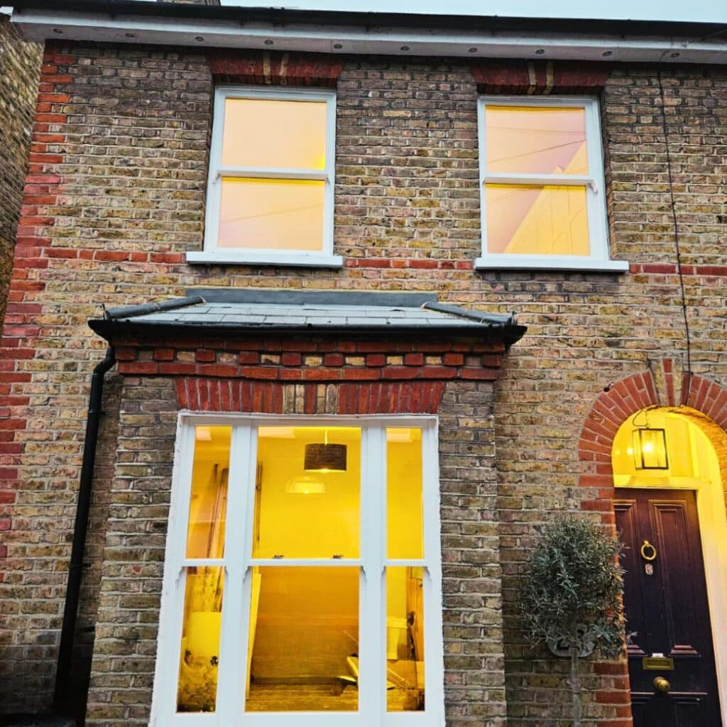 Conservation sash windows in London showcases four wooden windows, two on each floor. The lower ones emit a warm glow. The entrance features a black door with a curved top, illuminated by a glowing lantern above. An olive tree is planted in a pot by the door.