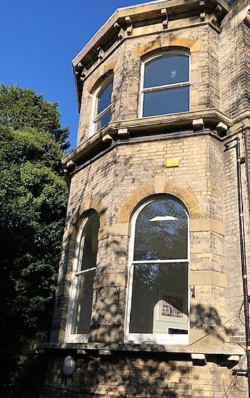 A historic building's corner in Cambridge showcases tall, arched sash windows framed by light brickwork. Sunlight casts shadows on the walls, while a large tree stands nearby against the clear blue sky.