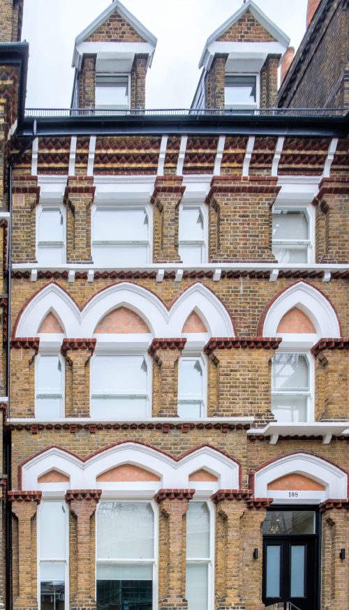 A Victorian-style brick building in North London boasts sash windows with arched white frames and decorative brick patterns. The facade features symmetrical design elements, complemented by charming dormer windows at the roof level.
